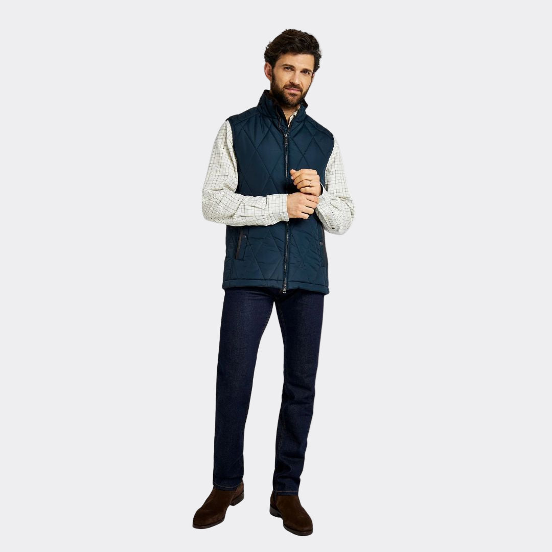 Man wearing a navy puffer vest over a white shirt with dark jeans on a white background