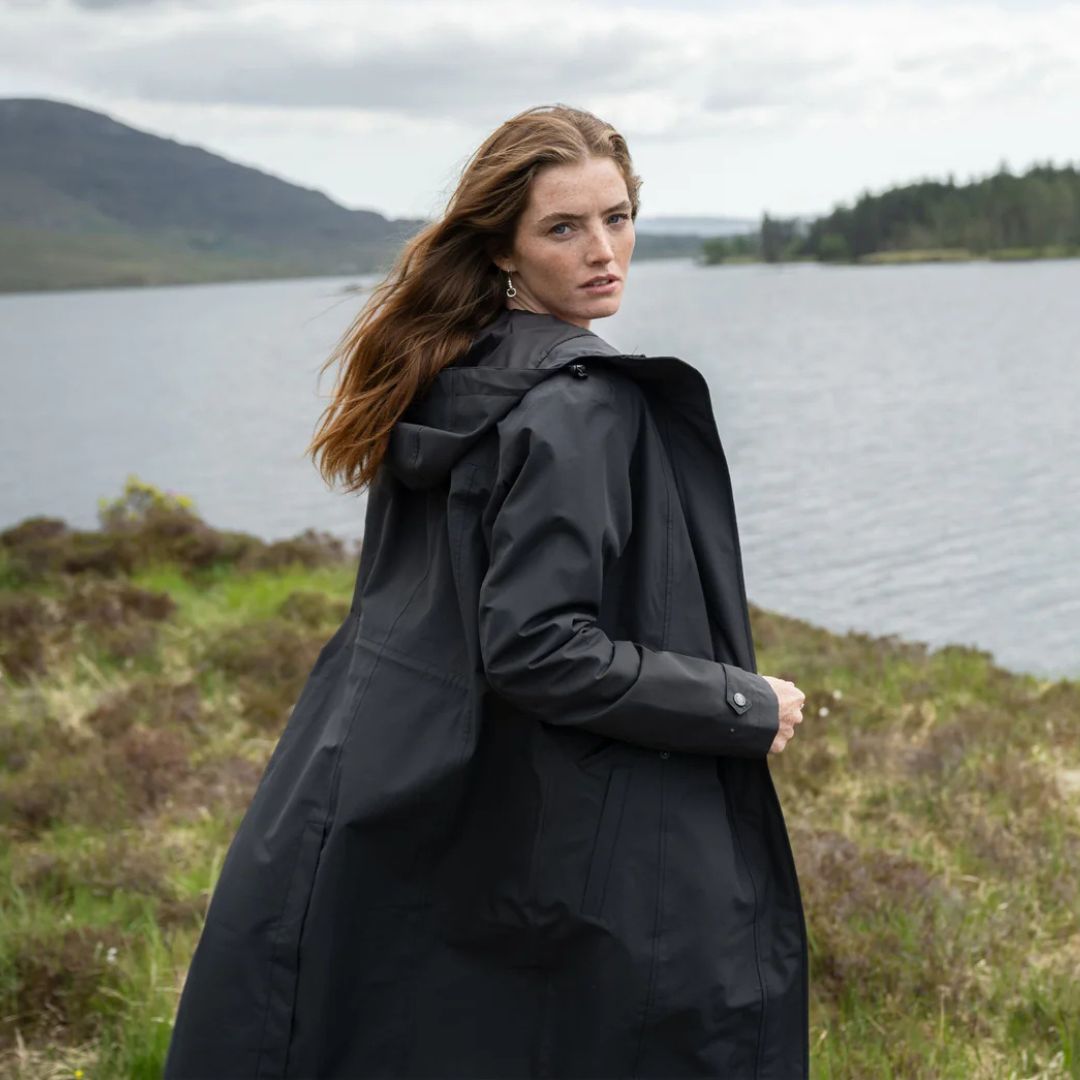 Woman wearing a black raincoat standing by a lake with mountains in the background