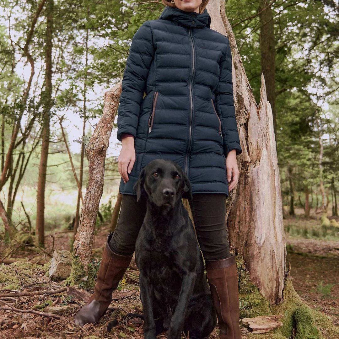 Woman in a blue puffer coat standing with a black dog in a forest setting