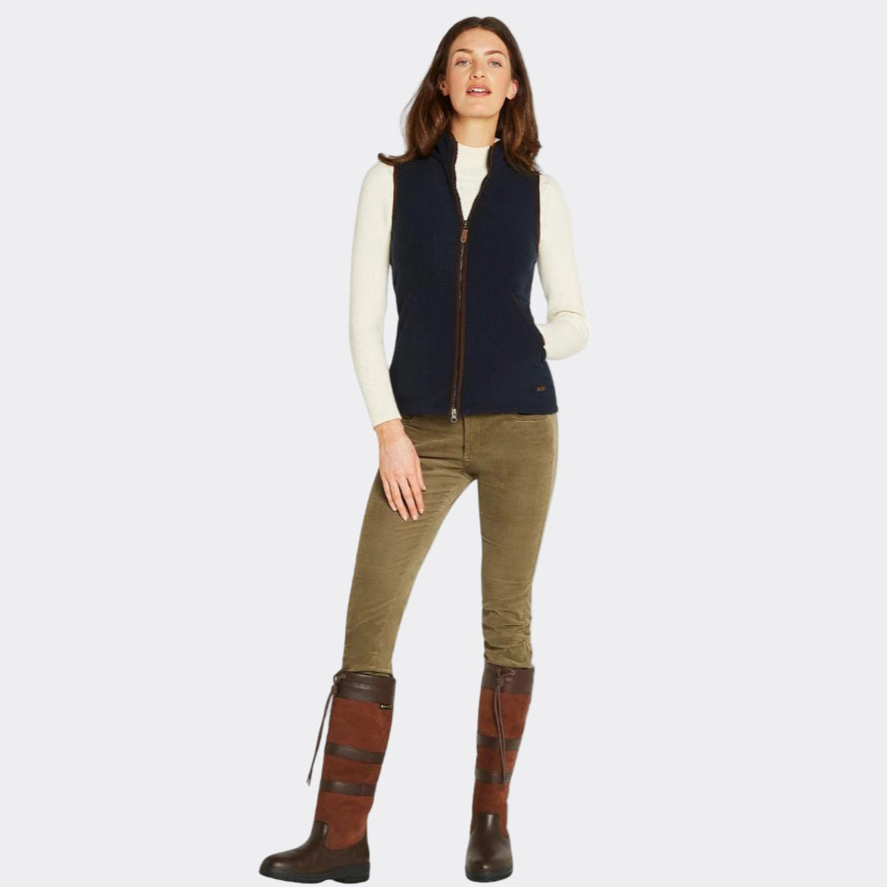 Woman wearing a navy vest, white shirt, green pants, and brown riding boots on a white background