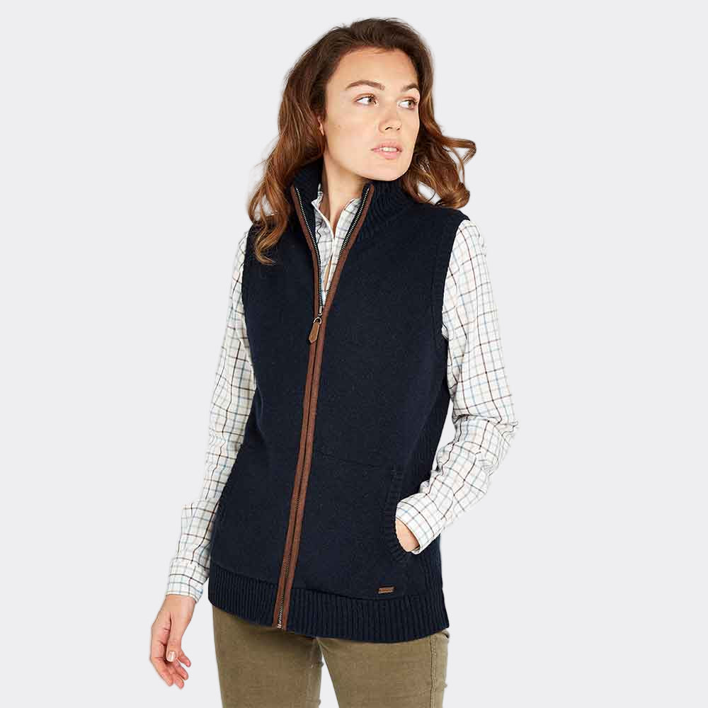 A woman wearing a navy-colored Dubarry Sheiddy Knit Gilet with a full zip, paired with a checkered shirt and knitwear. The gilet has soft, faux suede piping on the center front zip and inner neck tape.