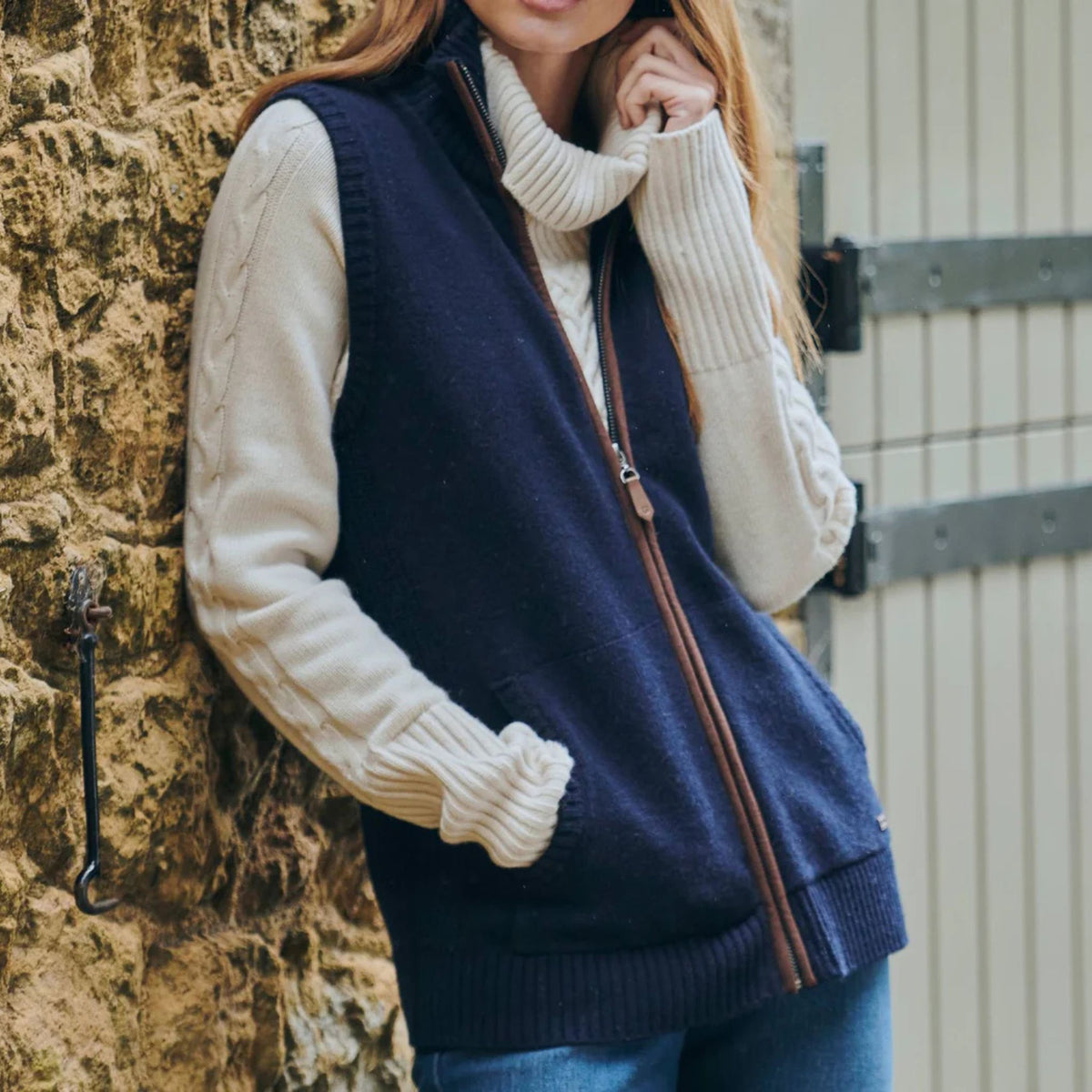 Person wearing a navy vest over a cream sweater against a stone wall.
