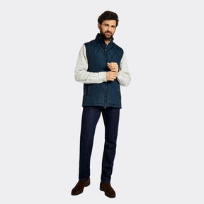 Man wearing a navy puffer vest over a white shirt with dark jeans on a white background