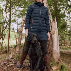 Woman in a blue puffer coat standing with a black dog in a forest setting